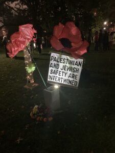 Outside in the dark at night, two large paper flowers are lit up with lights to demonstrate grief. A sign says: Palestinian and Jewish safety are intertwined.